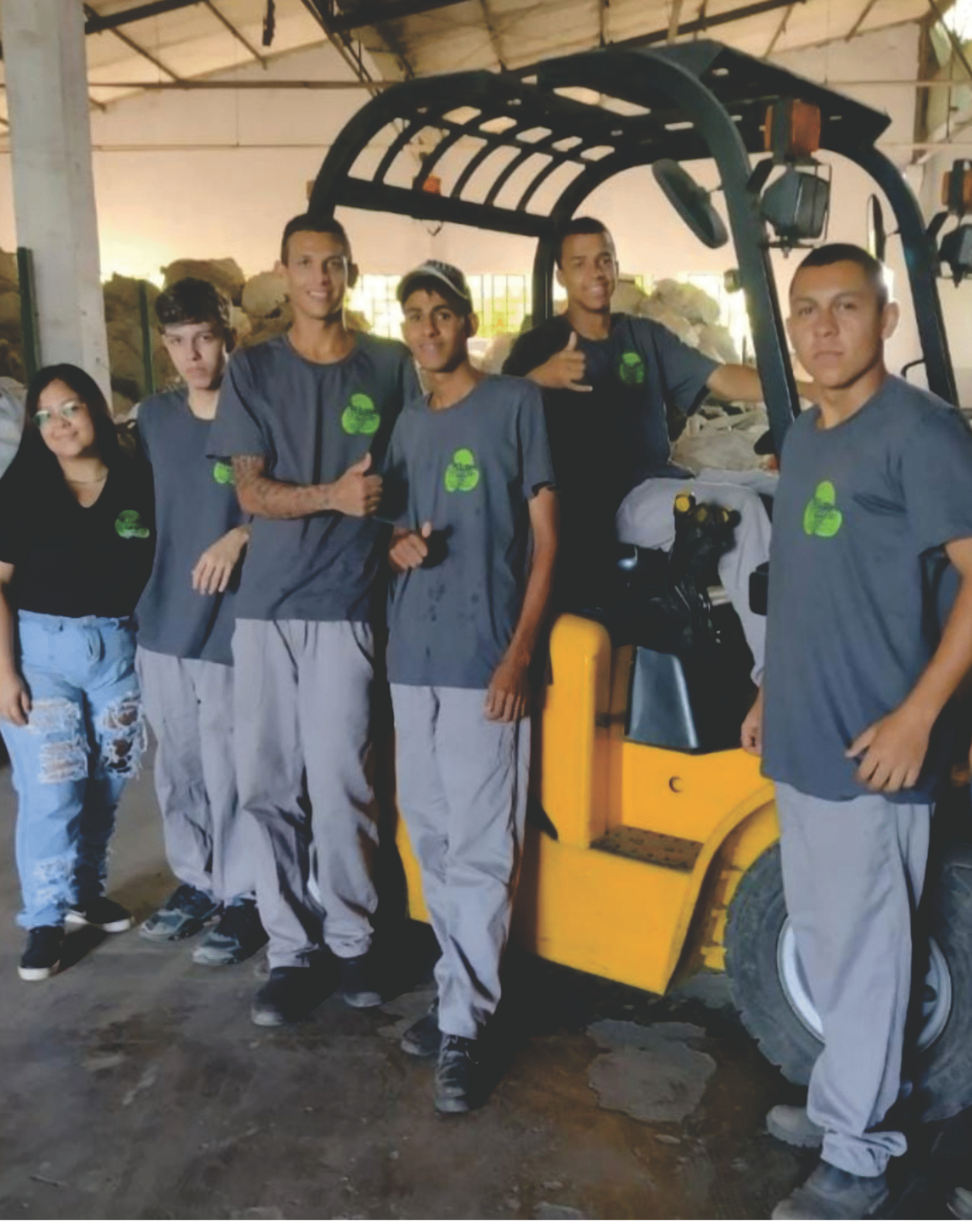 Grupo de profissionais em um galpão industrial vestindo camisetas personalizadas cinza com logotipo verde, posando ao lado de uma empilhadeira amarela.