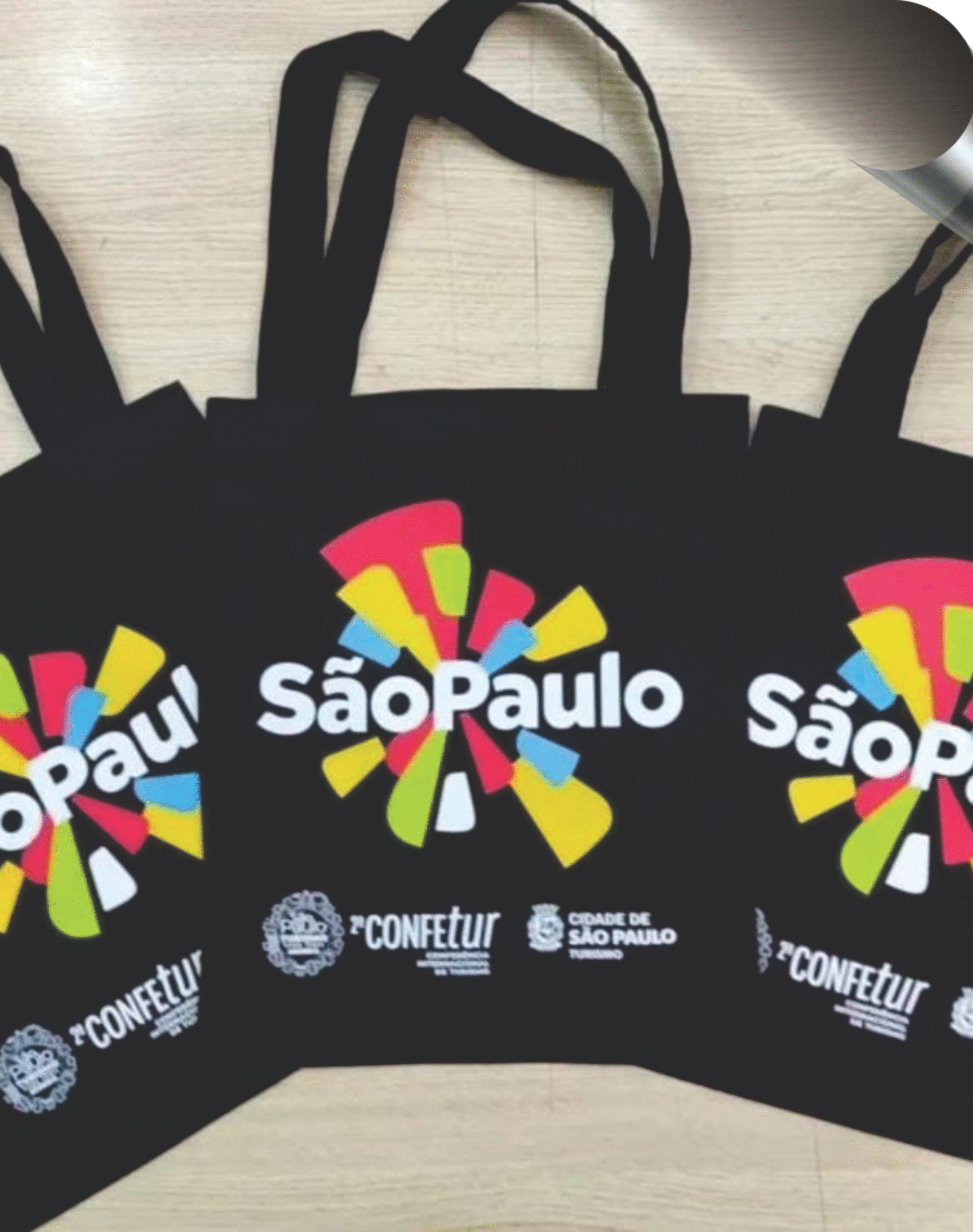 Trio de ecobags pretas personalizadas com estampa colorida da "2ª CONFETUR - Cidade de São Paulo", demonstrando o serviço de brindes personalizados da Maximus Print.