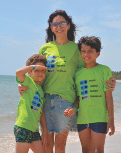 Mulher e duas crianças na praia vestindo camisetas personalizadas verde limão com estampas de interruptores indicando "Modo Férias ON" e "Trabalho OFF", produzidas pela Maximus Print.