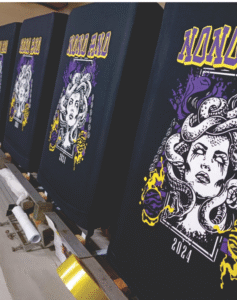 Linha de produção de camisetas pretas de formatura do 9º ano com estampa detalhada da Medusa em tons de roxo e dourado, produzidas pela Maximus Print.