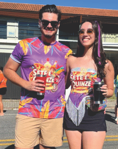 Casal vestindo abadás personalizados vibrantes em tons de roxo e laranja do bloco "Sete e Quinze", com estampas de pizza e bebidas, produzidos pela Maximus Print.